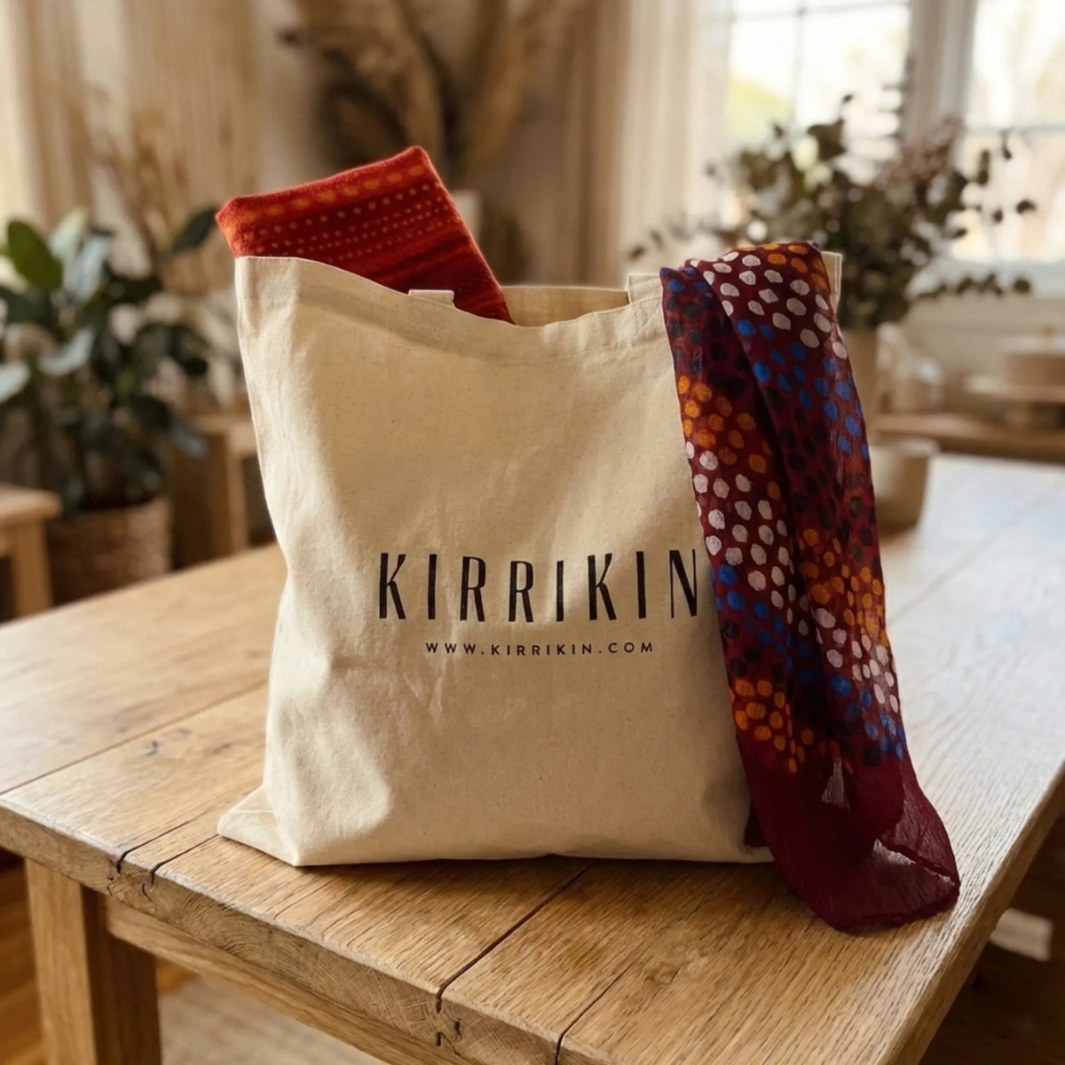 A beige canvas tote bag with the brand name 'KIRRIKIN' printed in black, sitting on a rustic wooden table. Two patterned scarves are draped from the top of the bag, one in rust red and orange, and the other in deep burgundy with orange, blue, and white geometric patterns.